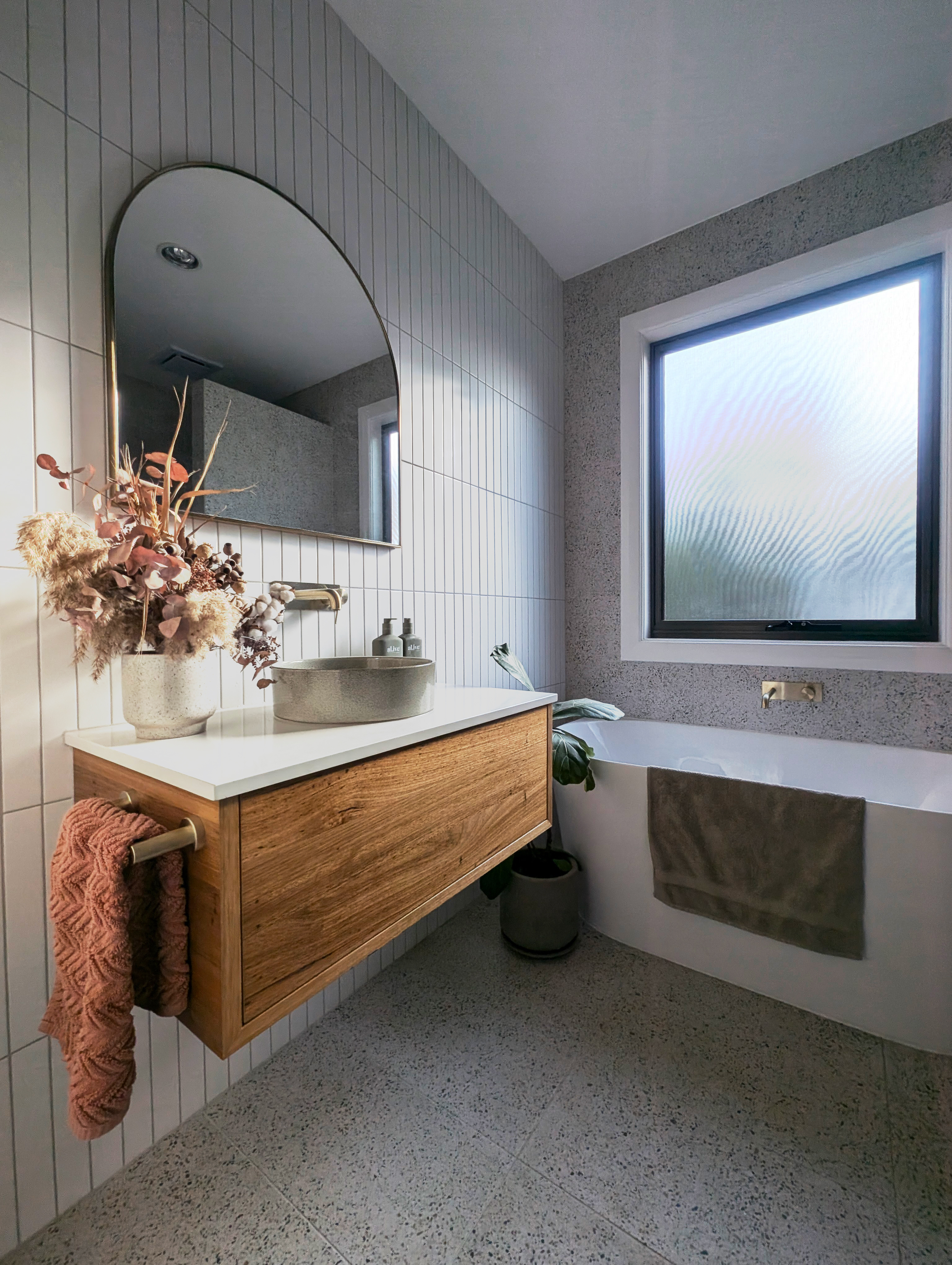 Cosy Balhannah bathroom rebuild — floating timber vanity with stone basin under an arched brass mirror, freestanding bath and heated terrazzo-look floors