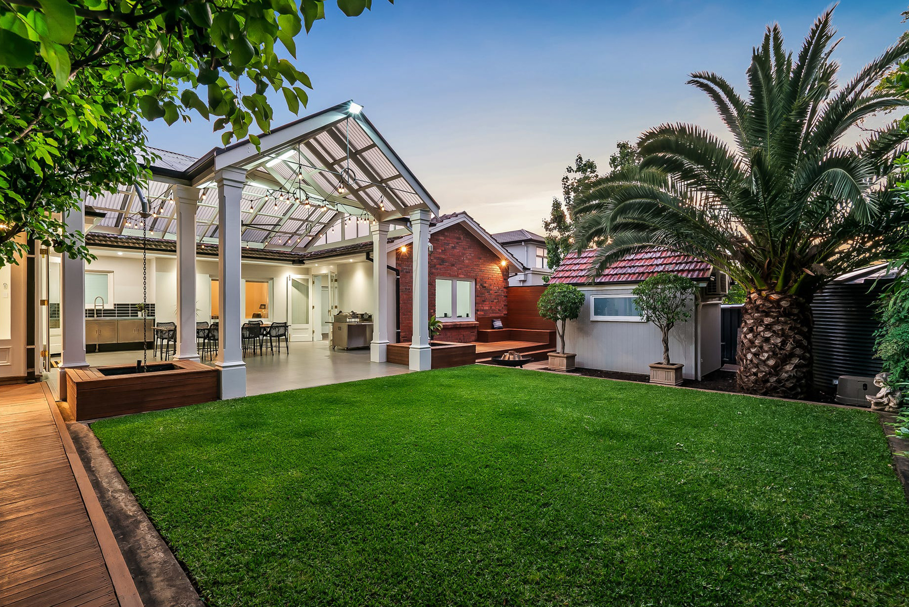 The Burnside outdoor living build at dusk — gabled pavilion with festoon lighting over the outdoor kitchen and dining, new decking and fire pit beside the red-brick home, studio build behind