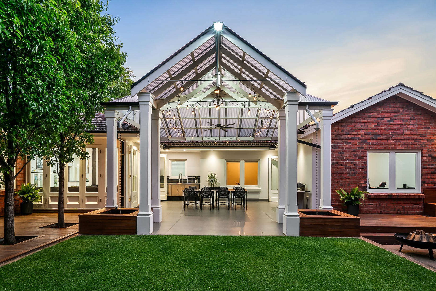 The Burnside pavilion from the lawn — gabled roof and festoon lights over outdoor dining, timber planter boxes with chain downpipes either side