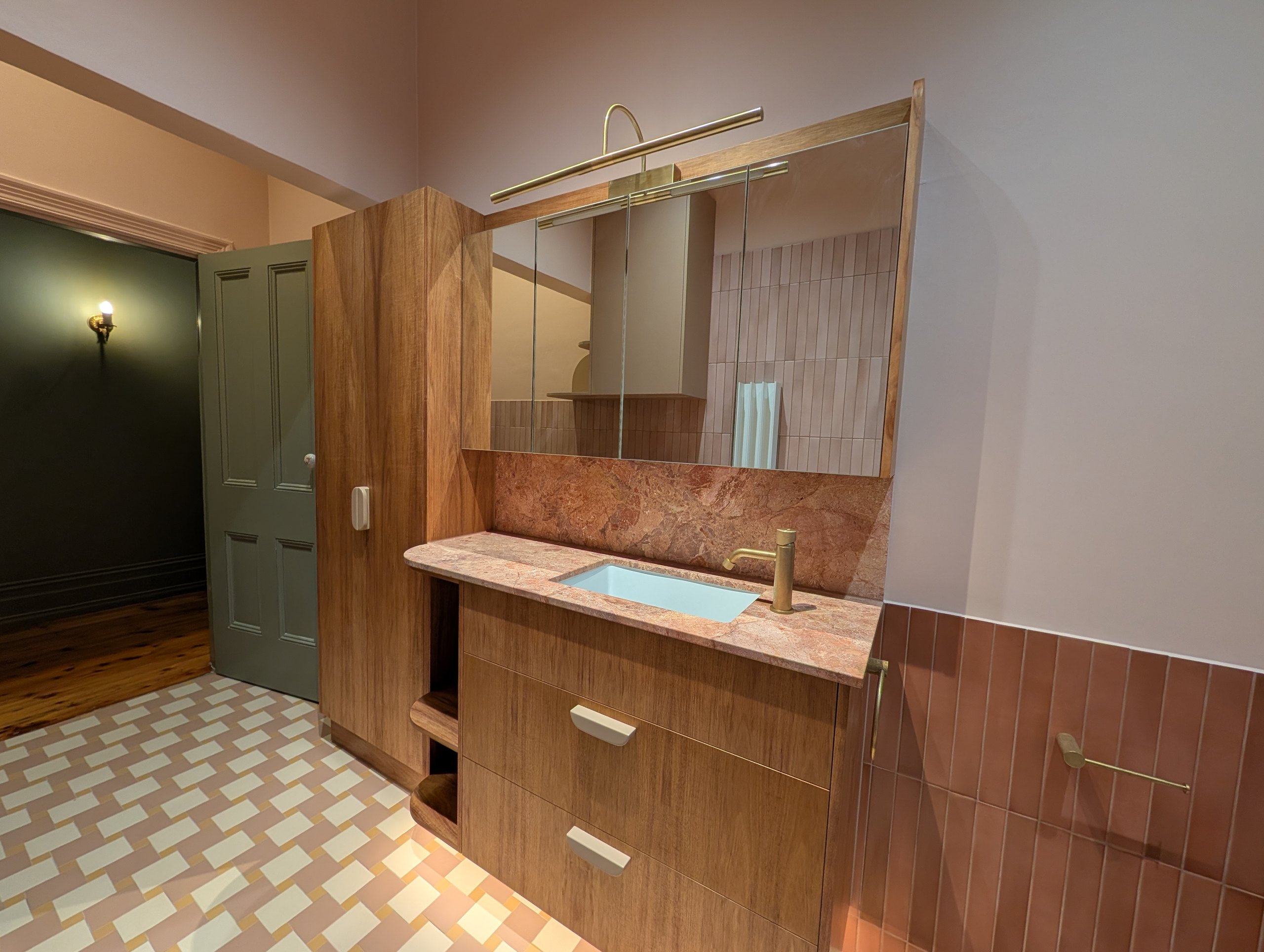 Bathroom vanity in timber with a stone top, mirrored cabinet and brass wall light, walk-in shower beyond