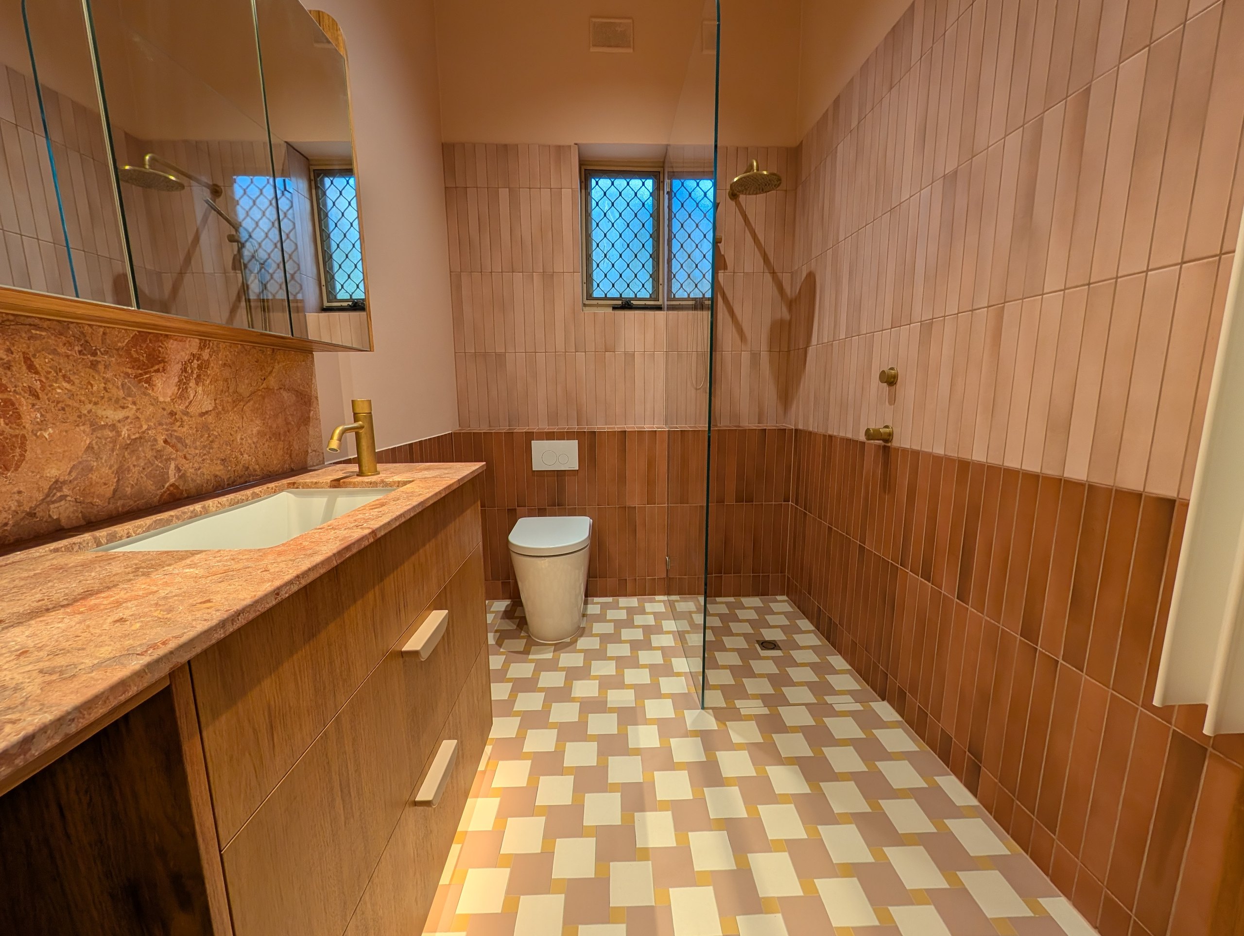 Pink tiled bathroom with a stone-topped timber vanity, living brass tapware, in-wall toilet and pinwheel floor