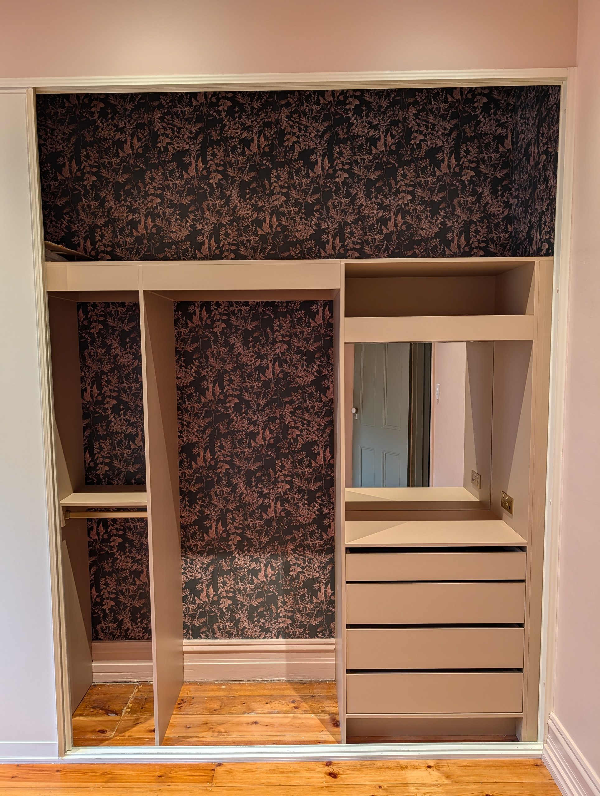 Bedroom robe in a soft pink panel colour with dark floral wallpaper behind the shelves and hanging space