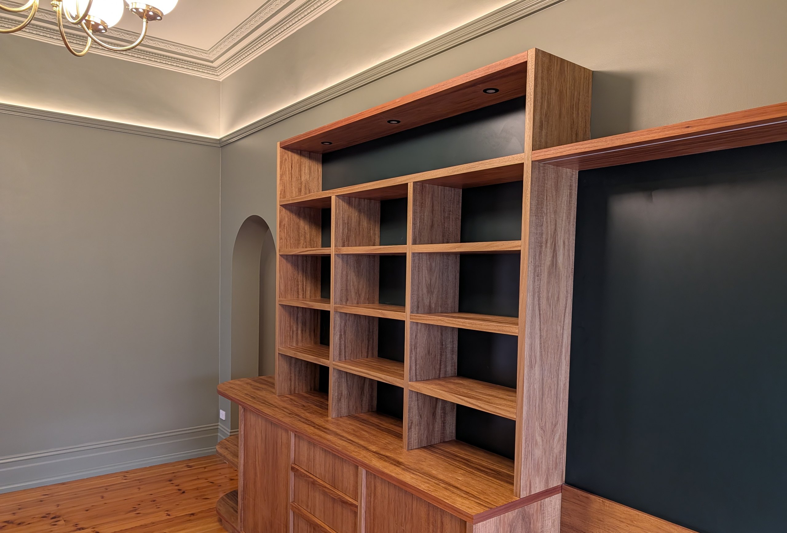 Timber bookshelf with downlights over the shelves, against green walls and a lit picture rail