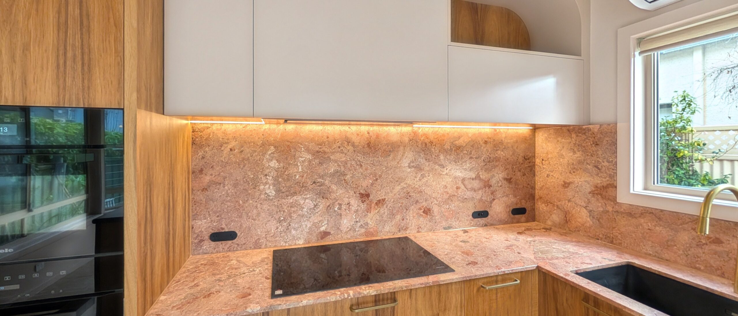 Timber and stone kitchen in the Glenelg cottage with under-cabinet LED lighting washing the stone splashback, stacked ovens on the left and the curved niche above