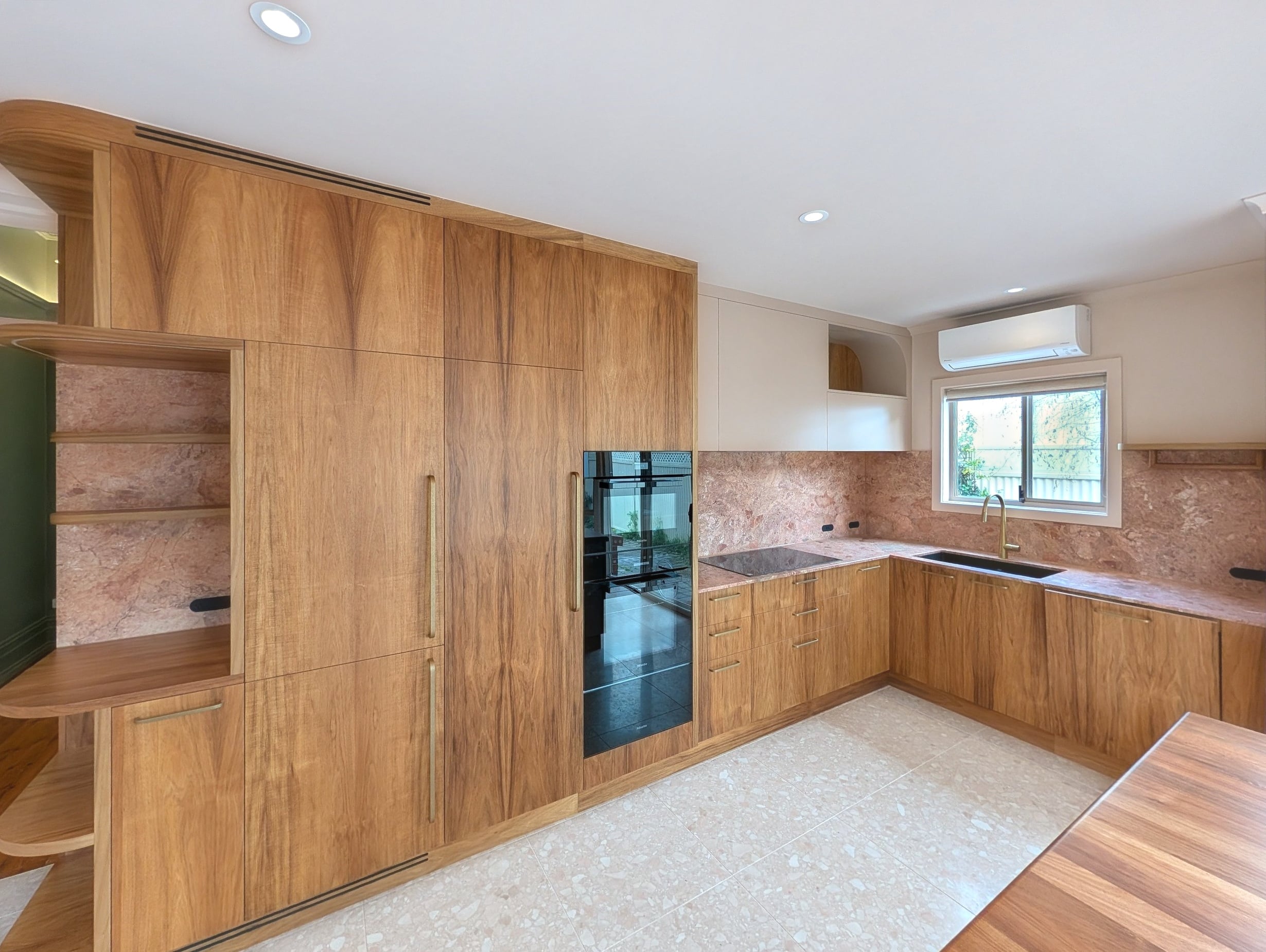 Timber-grain kitchen in the Glenelg cottage with integrated appliances, stone benchtops and splashback, and the curved open niche, seen from the dining end