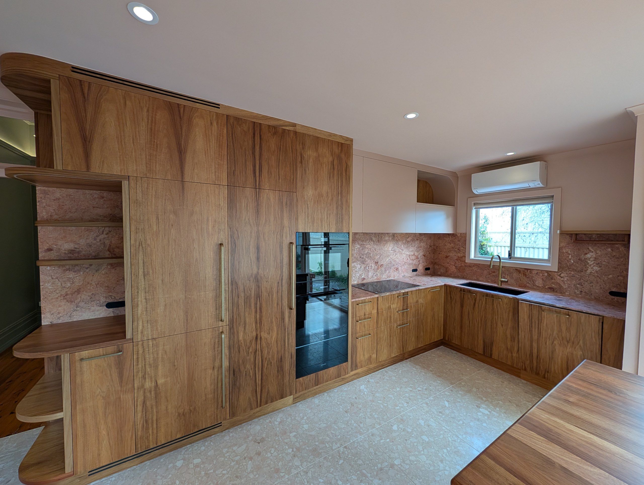 Timber-grain kitchen with integrated fridge, stacked ovens, stone benchtops and splashback, and a curved open niche