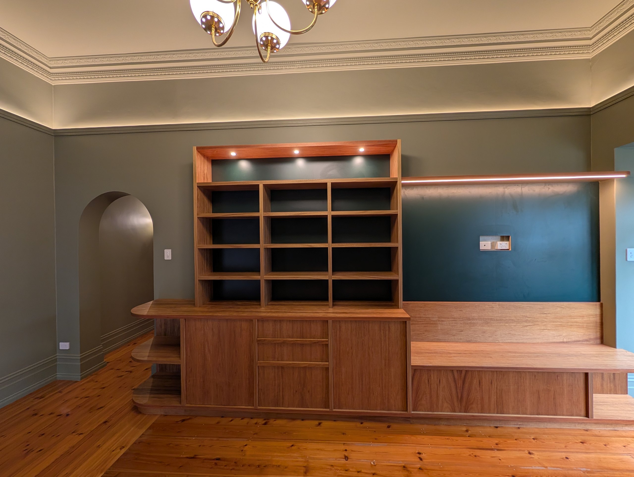 Living room wall of timber joinery in the Glenelg cottage: lit bookshelf, cabinets, bench seat and an LED shelf above the television wall