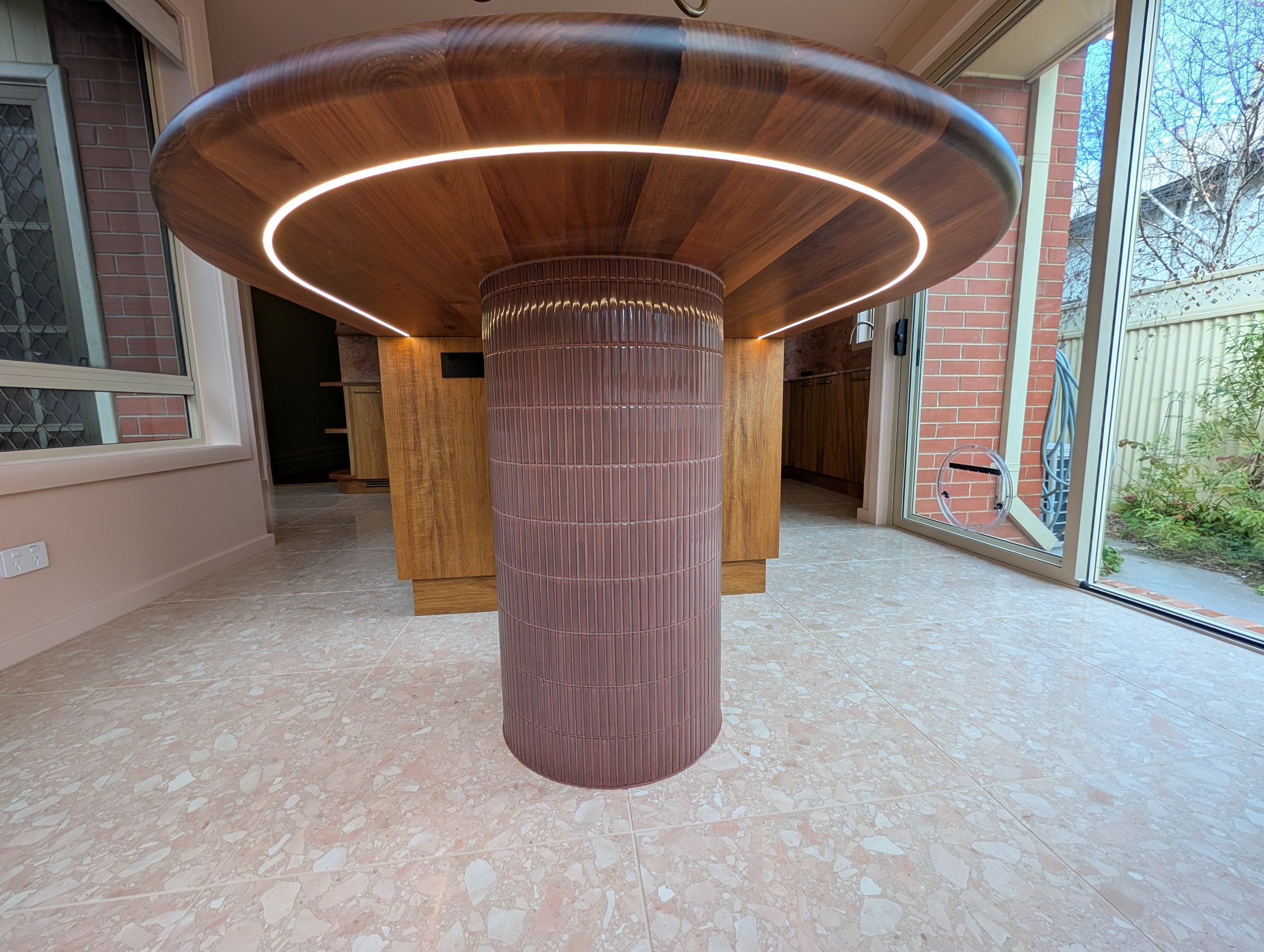 Underside of the rounded timber table showing the recessed LED ring, carried on a tiled pillar